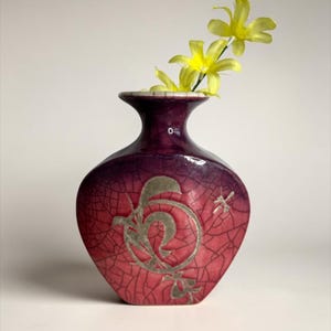 May include: A decorative vase with a deep purple and red crackle glaze finish. The vase features a silver floral design and holds yellow flowers. The vase is approximately 6 inches tall.