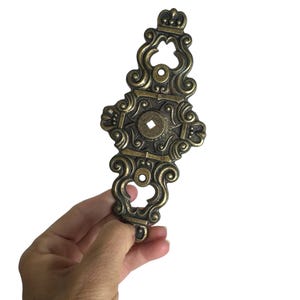 May include: Ornate antique brass furniture hardware. The decorative piece features a detailed, symmetrical design with scrollwork, a central diamond-shaped element, and a crown-like top. The hardware is held against a white background.