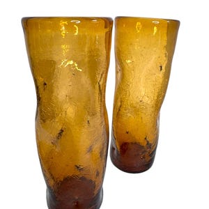 May include: Two amber-colored drinking glasses with a textured, crackled appearance. The glasses have a flared top and a rounded base, with a darker brown hue at the bottom. The glassware is likely vintage or handcrafted.