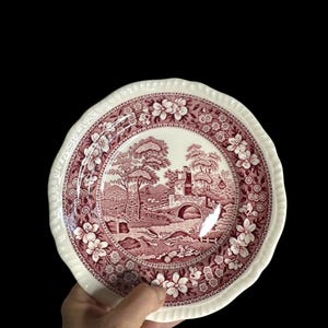 May include: A white and maroon decorative plate with a landscape scene in the center. The plate has a scalloped edge and a floral border. The landscape scene includes trees, a bridge, and a building.