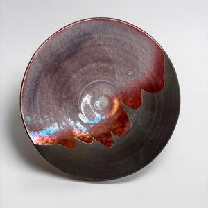 May include: A handmade ceramic bowl with a dark gray exterior and a deep burgundy interior. The rim has a scalloped edge with a vibrant, iridescent finish. The bowl's interior has a spiral design.