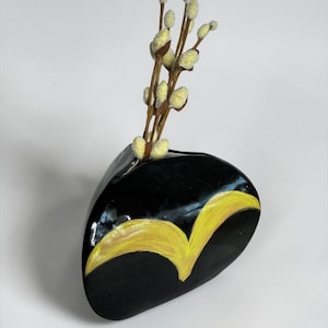 May include: A black, pebble-shaped vase with a yellow painted design, holding several stems of pussy willow. The vase is a unique decorative item, perfect for displaying small floral arrangements.