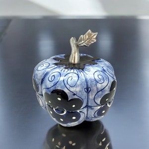 May include: A ceramic pumpkin-shaped trinket box with a blue and white floral design. The box has a silver-colored stem and leaf handle. The pumpkin has a glossy finish and is sitting on a reflective surface.