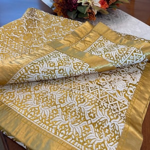 May include: A folded gold and white patterned tablecloth with a gold border. The tablecloth has a repeating design of white shapes on a gold background. The border is a solid gold color. The tablecloth is on a wooden table.