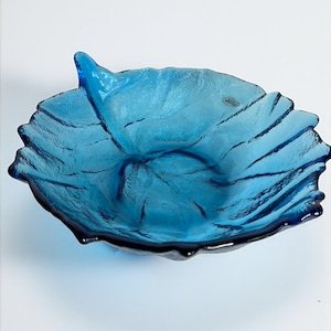 May include: A deep blue glass bowl shaped like a large leaf with a textured, organic surface. The bowl features a prominent stem-like protrusion on one side and a small, circular sticker near the rim, mimicking the veins and form of a natural leaf.