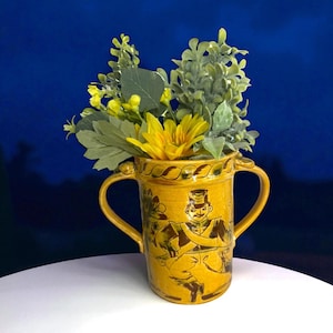 May include: A yellow ceramic vase with two handles, featuring a painted illustration of a man in a military uniform. The vase is filled with artificial yellow flowers and green foliage. The vase is on a white surface against a blue background.