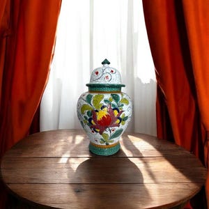May include: A decorative ceramic jar with a lid, featuring a floral design in red, yellow, green, and blue on a white background. The jar has a green base and rim, and is displayed on a round wooden table. The background includes orange curtains and a white curtain.