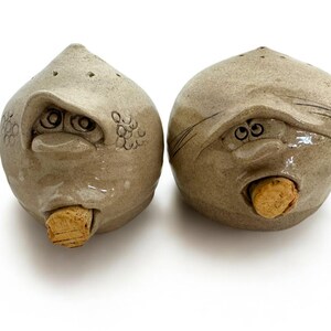 May include: Two whimsical, ceramic salt and pepper shakers. Each shaker is a light brown, rounded shape with a face carved into it. The faces have expressive eyes, noses, and mouths, with a cork in the mouth. The top of each shaker has small holes.