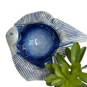 May include: A ceramic fish-shaped dish with a deep blue interior and white exterior. The dish has a textured surface with vertical lines on the fins and tail. The fish's head is white with a small eye. A green plant is in the foreground.