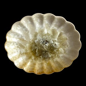 May include: An oval-shaped, off-white decorative bowl with a scalloped edge. The bowl's interior features a crystallized pattern with yellow and brown hues, creating a textured appearance. The bowl is set against a black background.