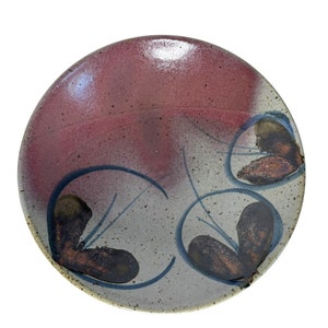 May include: A round ceramic plate with a red, white, and brown glaze. The plate has a design of three stylized leaves in brown with blue outlines.