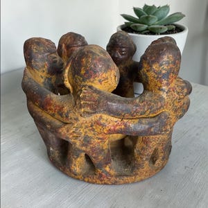 May include: A handcrafted, circular sculpture featuring six figures embracing. The figures are painted in shades of brown, red, and yellow, with visible texture. The sculpture has a rustic, aged appearance, suggesting it may be vintage or antique.