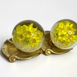May include: A pair of spherical resin paperweights or decorative ornaments featuring embedded bright yellow flowers and clear crystalline particles, resting on a textured, gold-toned metal base with a shell-like, scalloped design.