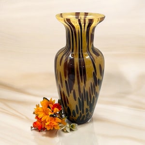 May include: A decorative glass vase with a flared top and a speckled pattern in shades of brown and amber. The vase is tall and slender, with a rounded base. A small bouquet of orange and white flowers sits beside the vase.