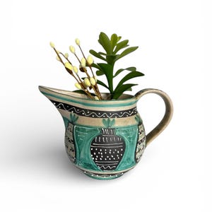 May include: A ceramic pitcher with a handle, painted with teal, black, and white designs. The pitcher holds a small green plant and yellow-tipped stems. The pitcher has a spout for pouring.