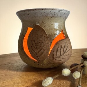 May include: A handcrafted ceramic candle holder with leaf cutouts. The holder is a mix of brown and tan with an orange glow emanating from within. The leaf designs are etched into the surface. A sprig of pussy willow is in the foreground.