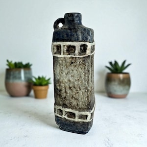 May include: A textured, ceramic bottle with a dark blue top and handle. The body is a mottled gray with white rectangular accents. The bottle is displayed in front of small potted plants, suggesting a decorative or home decor theme.