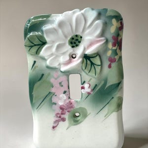 May include: A ceramic light switch cover with a floral design. The cover is painted with a large white flower with pink accents, green leaves, and pink and yellow floral accents. The background is a light green.