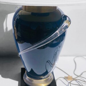 Puede incluir: Una l&aacute;mpara de mesa de vidrio azul con una pantalla blanca y una base dorada. La l&aacute;mpara tiene un dise&ntilde;o &uacute;nico con un remolino de vidrio transparente alrededor de la base.