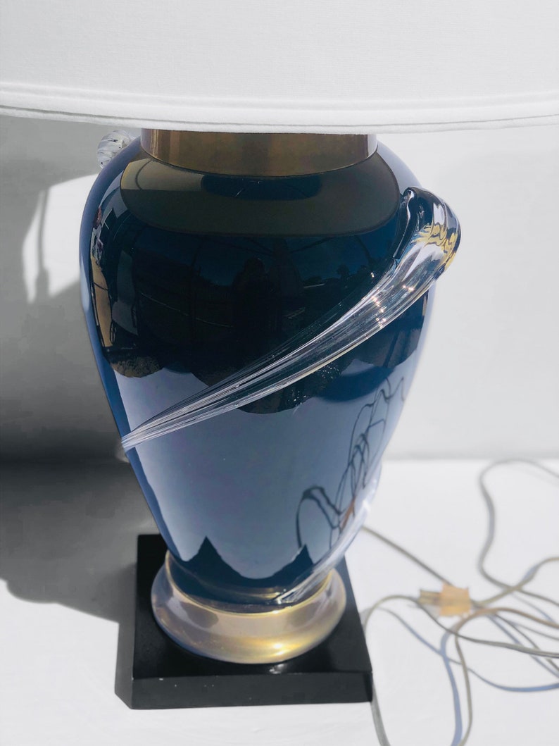 Puede incluir: Una l&aacute;mpara de mesa de vidrio azul con una pantalla blanca y una base dorada. La l&aacute;mpara tiene un dise&ntilde;o &uacute;nico con un remolino de vidrio transparente alrededor de la base.