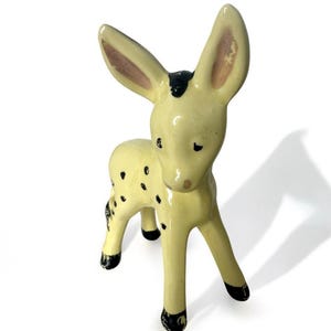 May include: A yellow ceramic figurine of a spotted deer with black hooves and a black spot on its head. The deer has pink ears and is standing on a white background.