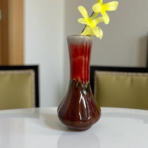 May include: A small, decorative vase with a flared top and a bulbous base. The vase has a gradient of colors, starting with a brown base, transitioning to red, and ending with a gray rim. Two yellow flowers are placed in the vase.