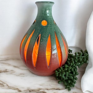 May include: A ceramic candle holder with a green and red gradient. The holder has a vase-like shape with cut-out designs that allow the warm orange glow of the candle inside to shine through. A small succulent plant is next to the candle holder.