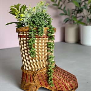 May include: A woven wicker boot-shaped planter with a brown and red design. The planter is filled with green artificial plants. The boot has a decorative woven strap around the ankle. The background is a soft pink.
