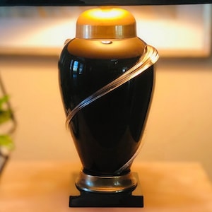 Puede incluir: L&aacute;mpara de mesa negra y dorada con un dise&ntilde;o &uacute;nico que presenta un acento de vidrio transparente curvado. La base de la l&aacute;mpara es una urna negra con una parte superior dorada y una banda dorada alrededor del medio.