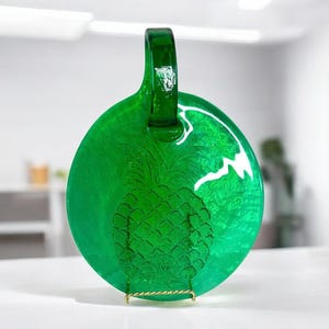 May include: A round, emerald green glass serving plate with a handle. The plate features an embossed pineapple design in the center. The plate is displayed on a gold-colored metal stand.
