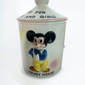 May include: A vintage ceramic jar with a lid, featuring a 3D Mickey Mouse figure. The jar is off-white with a pink rim and a lid with the text "FOR BOYS AND GIRLS". The words "Mickey Mouse" are printed at the bottom.