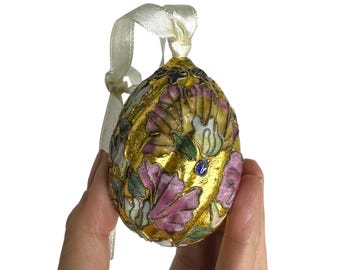 Könnte beinhalten: Ein eiförmiger Schmuckanhänger mit goldener Basis, verziert mit rosa und weißen Blumenmustern und grünen Blättern. Ein cremefarbenes Band ist zum Aufhängen befestigt. Der Anhänger hat einen kleinen blauen Edelstein und goldene Verzierungen.