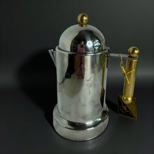 May include: A stainless steel coffee pot with a gold-colored handle and lid accents. The pot has a rounded body and a spout for pouring. A small tag with the text "VESSA" is attached to the handle.