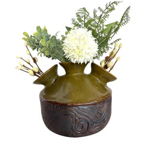 Puede incluir: Un jarr&oacute;n de cer&aacute;mica verde con una base marr&oacute;n texturizada y tres aberturas para flores. El jarr&oacute;n est&aacute; lleno de flores blancas y follaje verde.