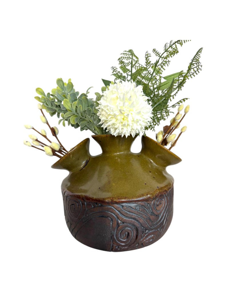 Puede incluir: Un jarr&oacute;n de cer&aacute;mica verde con una base marr&oacute;n texturizada y tres aberturas para flores. El jarr&oacute;n est&aacute; lleno de flores blancas y follaje verde.