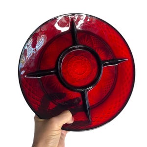 May include: A ruby red, divided serving dish with a textured surface and a central circular compartment. The dish has four black dividers extending from the center, creating separate sections. The dish is held by a hand against a white background.