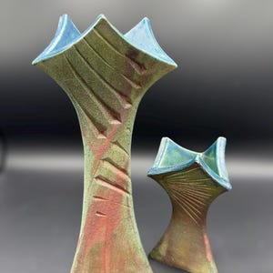 May include: Two ceramic vases with a unique, abstract design. The vases have a tall, slender shape with a flared top and a textured surface. The color palette includes shades of green, red, and blue, creating a striking visual contrast. The vases are handmade.