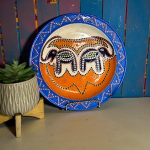 May include: A decorative ceramic plate with a blue rim and an orange center. The plate features a design of two elephants with black and white dot patterns. The plate is displayed on a gold metal stand.