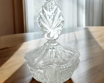 May include: A clear glass candy dish with a decorative, faceted lid. The lid has a large, ornate, crystal knob in the shape of a leaf.