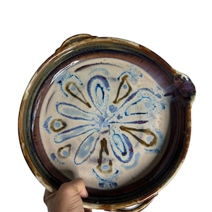 May include: A round ceramic serving dish with a floral design in shades of blue, brown, and white. The dish has a dark brown rim and two handles. The dish is held by a hand against a white background.