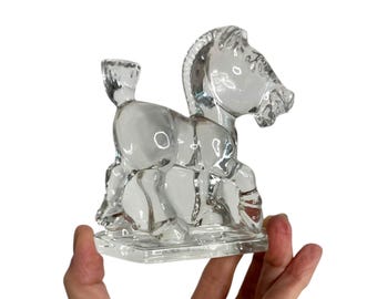 May include: Clear glass horse figurine with a candle holder on its back. The horse is stylized with a blocky design and sits on a square base. The glass is transparent, allowing light to pass through.