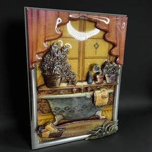 May include: A three-dimensional ceramic wall plaque featuring a bathroom scene with a clawfoot tub, potted plants on a windowsill, and a basket of items on the floor. The artwork is framed in a silver-colored metal border with a glossy finish.
