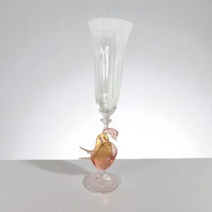 May include: A clear champagne flute with a pink and gold glass swan base. The flute has vertical ridges and a round base. The swan has a pink body and gold wings. The glass is transparent.
