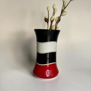 May include: A ceramic vase featuring horizontal bands of black, white crackle glaze, and red. The vase has a flared rim and base, and contains several pussy willow branches with fuzzy, light-colored buds extending from the top opening.