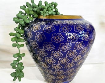 Puede incluir: Un jarrón de cerámica azul con borde dorado y estampado floral dorado. El jarrón está lleno de una planta suculenta verde. El jarrón mide aproximadamente 20 cm de alto y 15 cm de ancho.