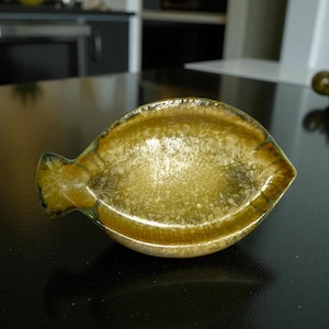 May include: A decorative, leaf-shaped dish with a textured, golden-yellow interior and a brown rim. The dish has a small, pointed handle on one side. The dish is likely made of ceramic or glass.