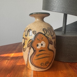 May include: A ceramic vase with a tan background and a cartoon face design in shades of brown and orange. The vase has a narrow neck and a wide base. The vase is approximately 7 inches tall.