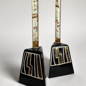 May include: A pair of decorative candle holders. Each holder has a black base with gold lightning bolt design and a clear, cylindrical stem filled with gold leaf flakes. The bases are outlined in white.