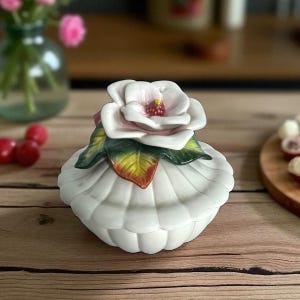 May include: A white ceramic trinket box with a flower-shaped lid. The lid features a detailed, light pink and white flower with green and yellow leaves. The box has a scalloped design and sits on a wooden surface.