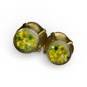 May include: A pair of vintage clip-on earrings. Each earring features a clear, round, glass-like orb filled with small yellow flowers and green accents. The orbs are set in ornate, gold-toned metal leaf-shaped settings.